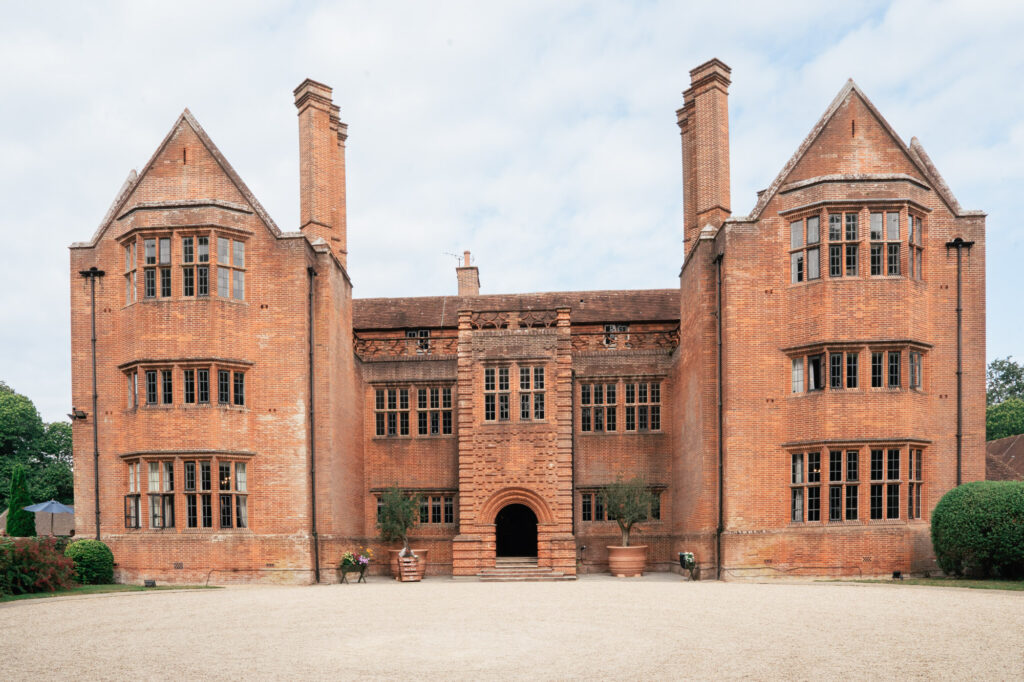 A large, symmetrical red-brick mansion with two tall chimneys, large sash windows, and an arched central entrance, set against a cloudy sky and surrounded by a gravel driveway.