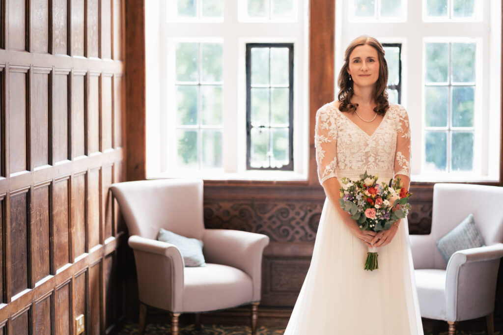A bride in a white wedding dress holding a bouquet stands between two armchairs in a bright room with wood paneling and large windows.
