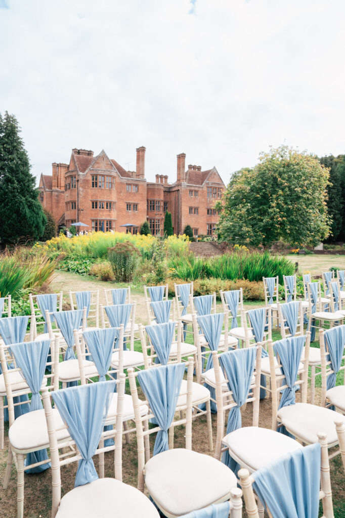 Rows of white chairs with light blue fabric draped over the backs are arranged outdoors on a lawn, facing a large, historic brick mansion with gardens and greenery in the background.