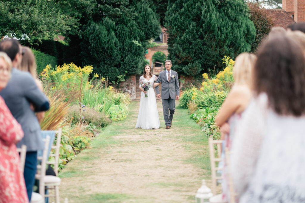 A bride in a white dress, holding a bouquet, walks down an outdoor aisle alongside an older man in a grey suit as guests seated on either side watch during a garden wedding ceremony.
