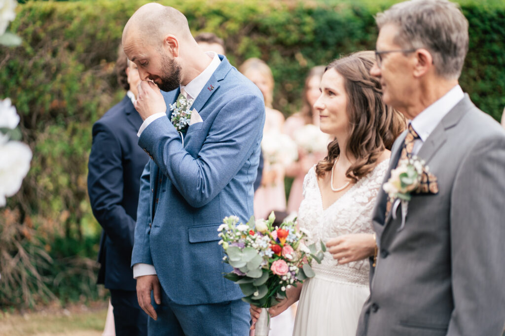A groom in a blue suit wipes his eyes emotionally while standing next to a smiling bride in a white dress holding a bouquet, with another man in a gray suit by their side outdoors.