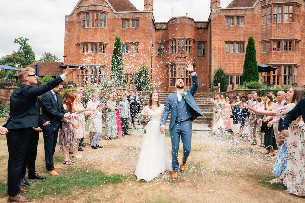 A bride and groom walk joyfully outside a large brick building, surrounded by guests throwing confetti in celebration. The couple smiles and holds hands, with friends and family cheering on both sides.