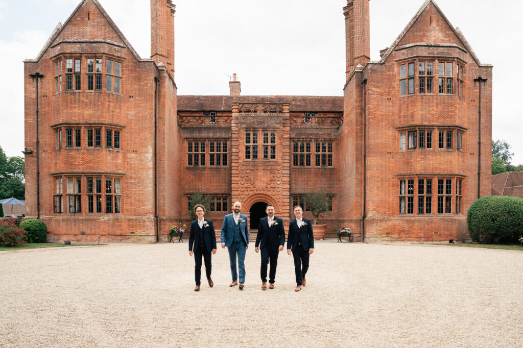 Four people dressed in formal suits walk together on a gravel path in front of a large, historic red-brick mansion with tall chimneys and many windows. The sky is overcast.