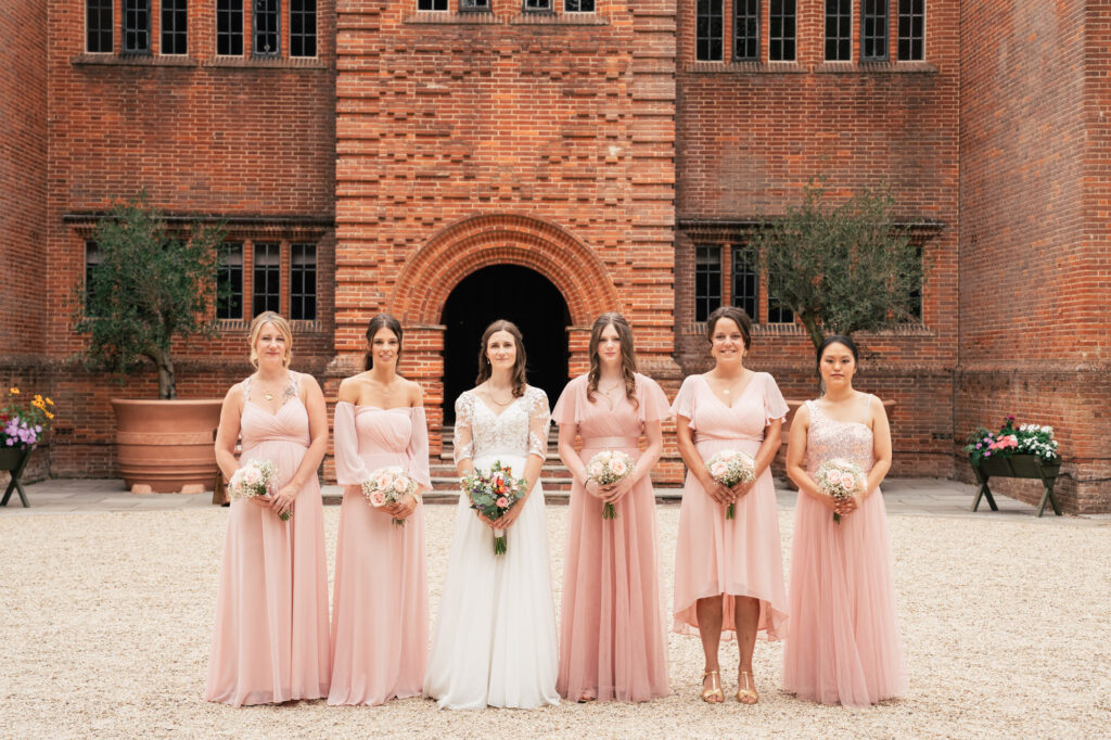 A bride in a white dress stands outside with five bridesmaids in matching blush pink dresses, all holding bouquets, in front of a brick building with arched entry and windows.