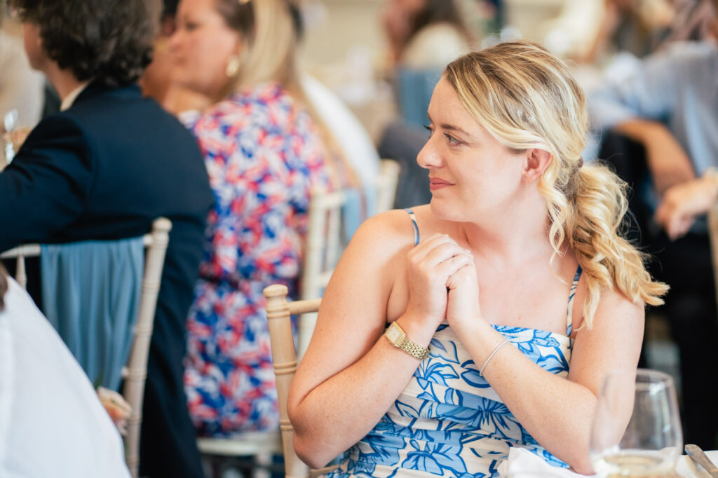 A woman in a blue and white floral dress sits at a table, smiling and looking to the side during what appears to be a social event. Other people are seated around her in the background.