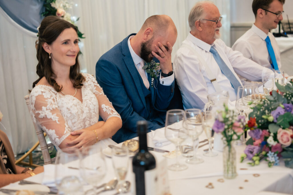 A bride in a lace dress smiles, sitting beside a groom in a blue suit who covers his face with his hand at a decorated wedding reception table with guests, flowers, and glasses.