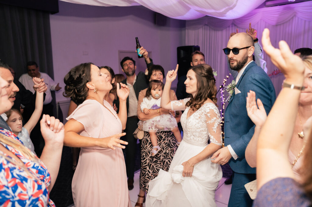 A group of people celebrate at a wedding reception. The bride in a white dress and groom in sunglasses are dancing, surrounded by smiling guests of all ages, including a child, under purple-tinted lights.