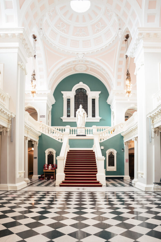 A grand hall with a black-and-white checkered floor, a red-carpeted staircase, ornate white railings, a statue at the top of the stairs, tall columns, and vaulted ceilings with chandeliers.