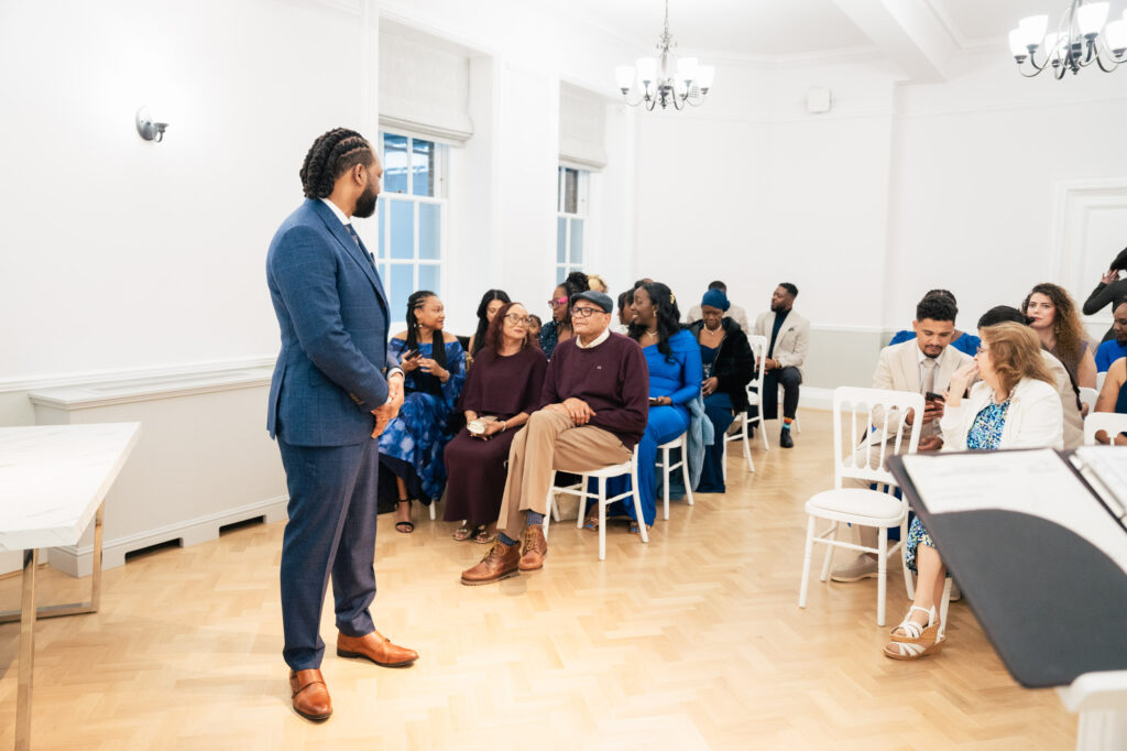 A man in a blue suit stands at the front of a bright room, facing a seated audience who are smiling and engaged. The room has wooden floors, white walls, and large windows.