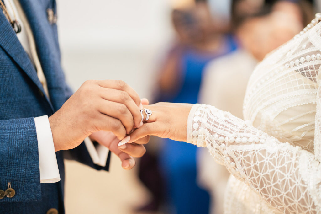 A person in a blue suit places a ring on the finger of another person wearing a white, intricately detailed dress, during a wedding ceremony. The background is softly blurred.