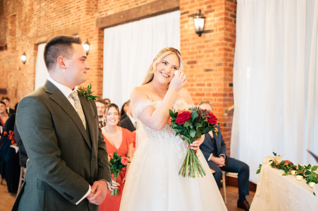 A bride in a white gown wipes a tear while holding a bouquet of red and white flowers, standing beside a smiling groom in a dark suit at an indoor wedding ceremony. Guests are seated in the background.