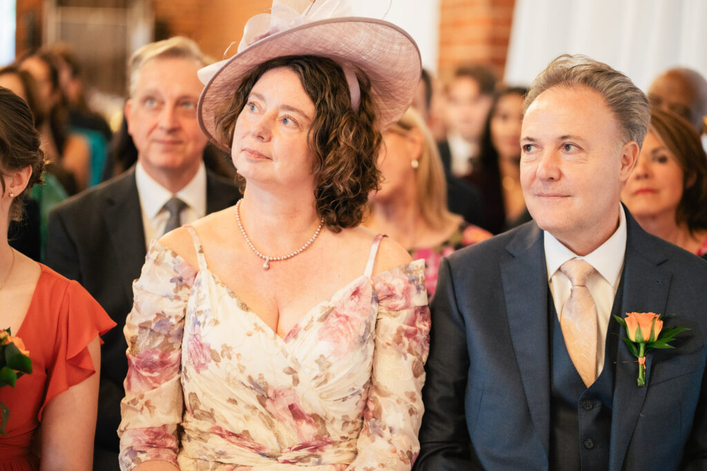 A woman in a floral dress and large hat sits beside a man in a suit and tie at a formal event, possibly a wedding, with other well-dressed people seated around them. Both look forward, appearing attentive.