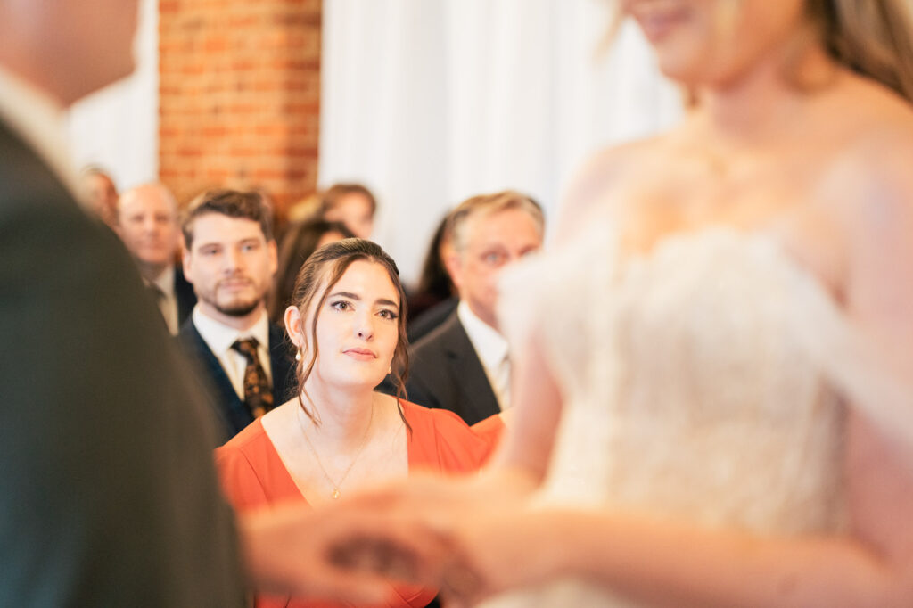 A woman in an orange dress looks on emotionally as a couple exchanges vows at a wedding ceremony, with other guests seated in the background.