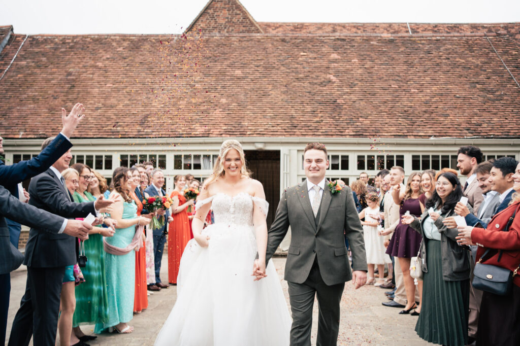 A bride and groom walk hand in hand outside, smiling, while guests line both sides, clapping and throwing confetti in celebration. Everyone is dressed formally in colorful attire. A rustic building is in the background.