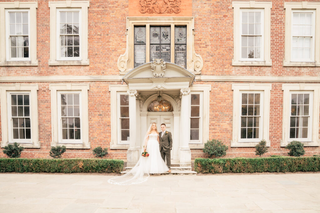 A bride and groom stand together in front of a large, historic brick building with tall windows and ornate door, posing for a wedding photo. The bride holds a bouquet and wears a long white dress and veil.