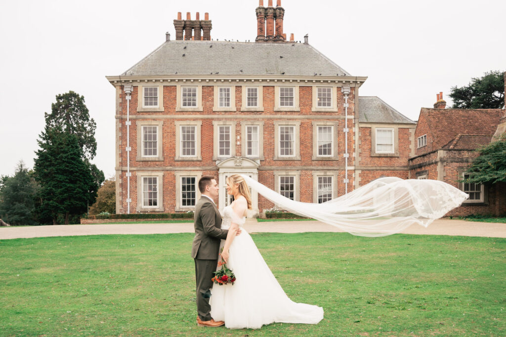 A bride and groom stand facing each other on a lawn in front of a large, elegant mansion; the bride’s veil flows dramatically in the wind as they embrace, and she holds a bouquet of flowers.