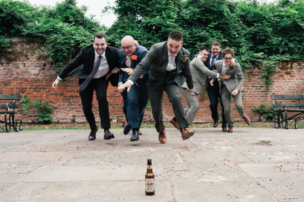 Six men in suits eagerly race toward a bottle placed on the ground outdoors, with a brick wall and greenery in the background. They appear playful and excited, mid-action as they lean forward with outstretched arms.