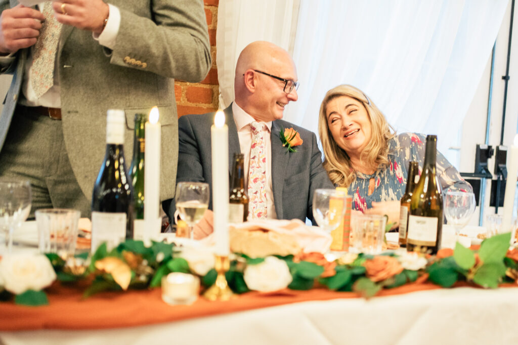 A smiling man and woman sit at a decorated table with candles, flowers, and wine bottles, enjoying a joyful moment at a formal event or celebration.