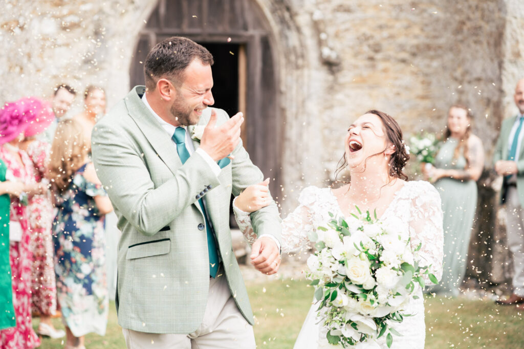 A bride and groom laugh joyfully as confetti is thrown around them outside a stone building, surrounded by smiling guests. The bride holds a white bouquet and wears a lace dress; the groom wears a green jacket and teal tie.
