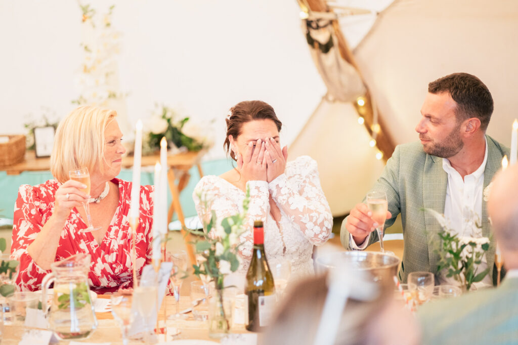 A bride in a white lace dress covers her face, laughing or crying, while seated between a woman in a red floral dress and a man in a green suit. They all hold champagne glasses at a decorated table.
