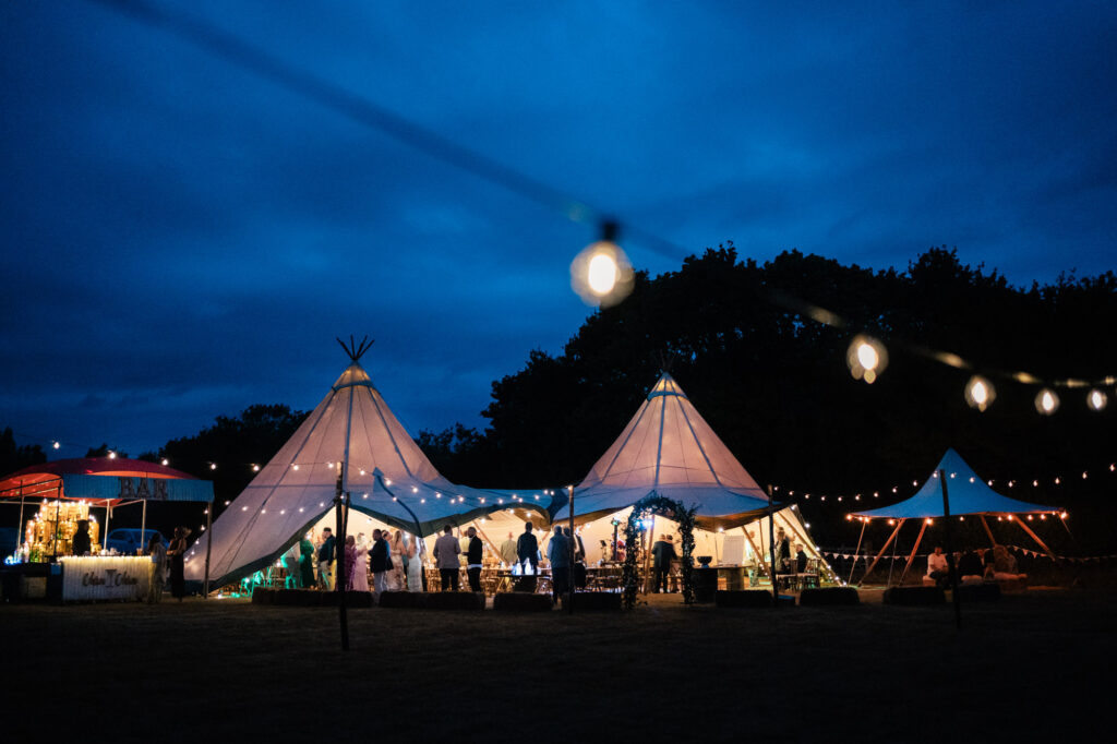 A nighttime outdoor event with illuminated tipi tents, people gathered inside, string lights overhead, and a dark blue sky in the background.