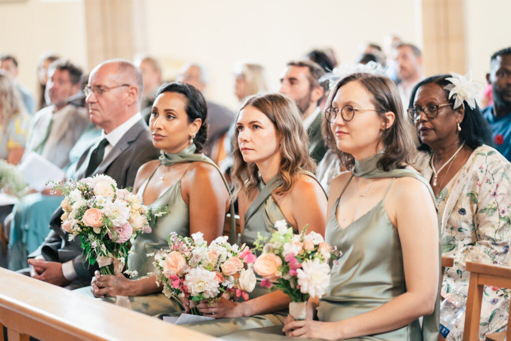 Three bridesmaids in matching sage green dresses sit in a church pew, holding bouquets of flowers. Other well-dressed guests are seated behind them, all facing forward and appearing attentive.