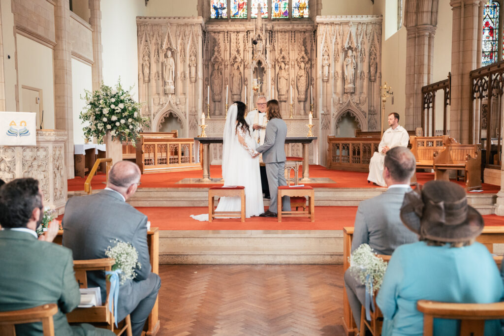 A bride and groom stand at the altar holding hands in a church, with a priest seated behind them. Guests are seated in pews, watching the wedding ceremony. The altar is ornate with detailed carvings and stained glass.