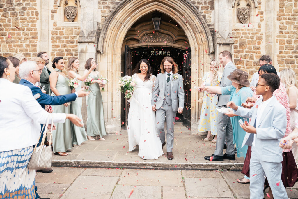 A bride and groom walk hand in hand outside a stone church as guests in colorful outfits throw flower petals in celebration. Bridesmaids in green dresses stand to the left. Everyone is smiling and joyful.