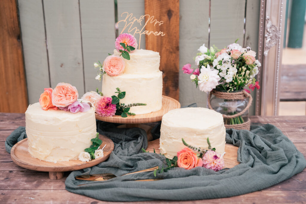 Three white frosted cakes decorated with fresh flowers sit on wooden stands, accompanied by a bouquet of flowers in a vase, all arranged on a wooden table with green fabric and gold cutlery.