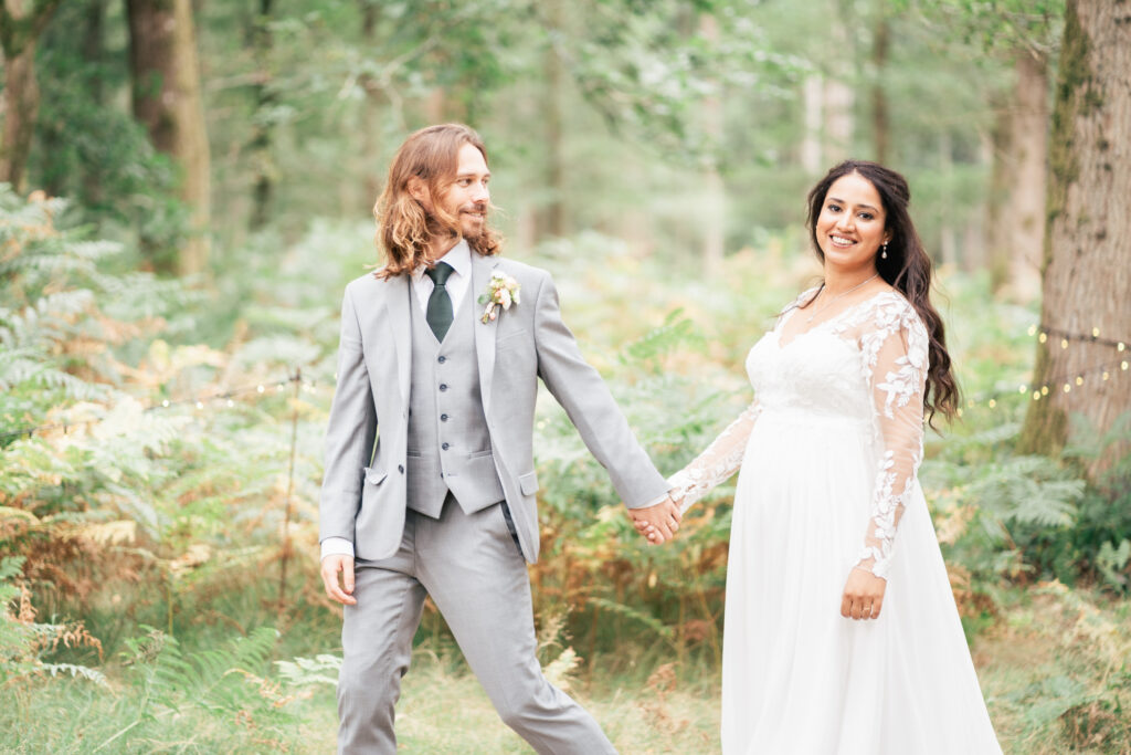 A bride in a white, long-sleeved lace dress and a groom in a light gray suit hold hands and smile while walking outdoors in a lush, green forest setting.