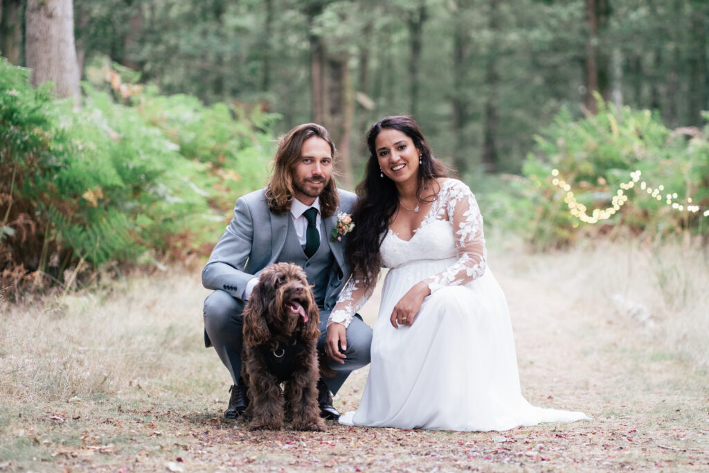 A bride and groom kneel on a forest path with greenery around them, smiling at the camera. The groom wears a gray suit, the bride wears a white gown, and a brown dog sits between them, its tongue out.