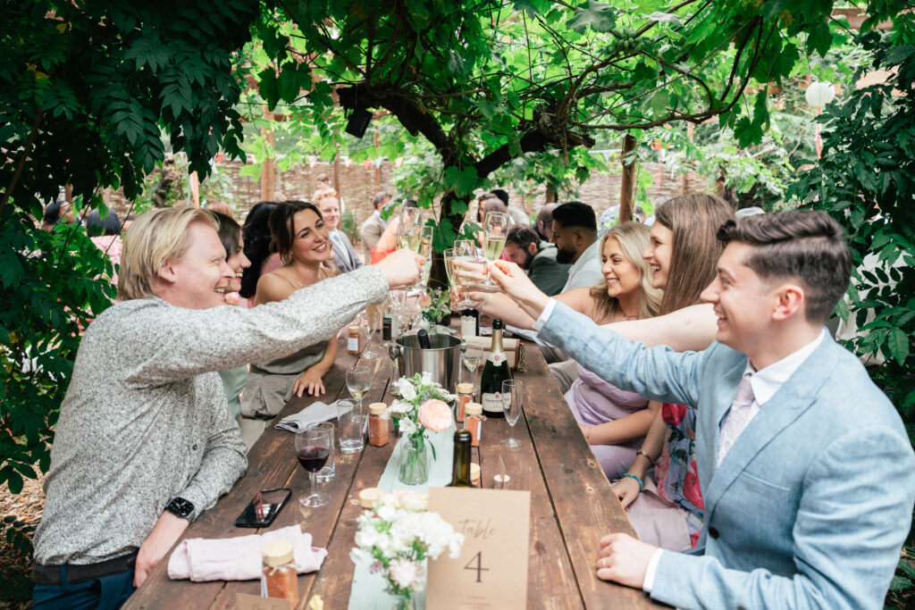 A group of people sit around a wooden outdoor table, raising glasses in a toast. They are smiling and surrounded by greenery and flowers, creating a festive, joyful atmosphere.