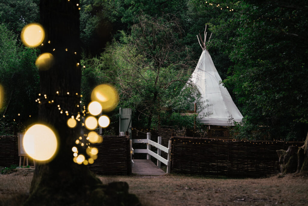 A white teepee stands in a wooded area, surrounded by trees and wooden fences. Warm string lights hang on tree trunks, creating a soft, glowing bokeh effect in the foreground.