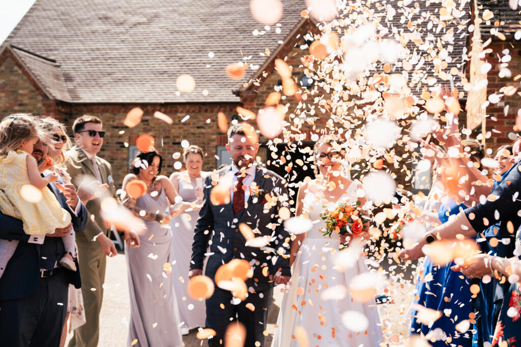 A bride and groom smile as guests shower them with confetti outdoors. Captured by a Surrey wedding photographer, bridesmaids and other guests, some holding children, celebrate around the happy couple in bright sunlight near rustic brick buildings.