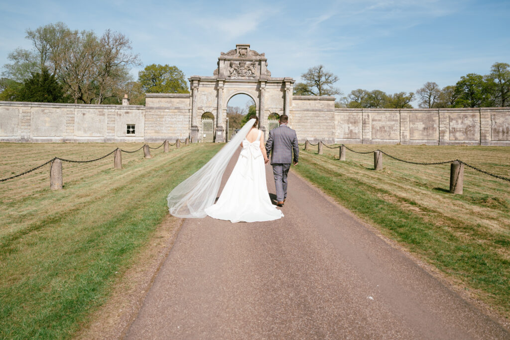 A bride and groom walk hand in hand down a path toward a grand stone archway, surrounded by green lawns and trees, under a blue sky. Captured by a Surrey wedding photographer, the bride’s veil flows behind her.