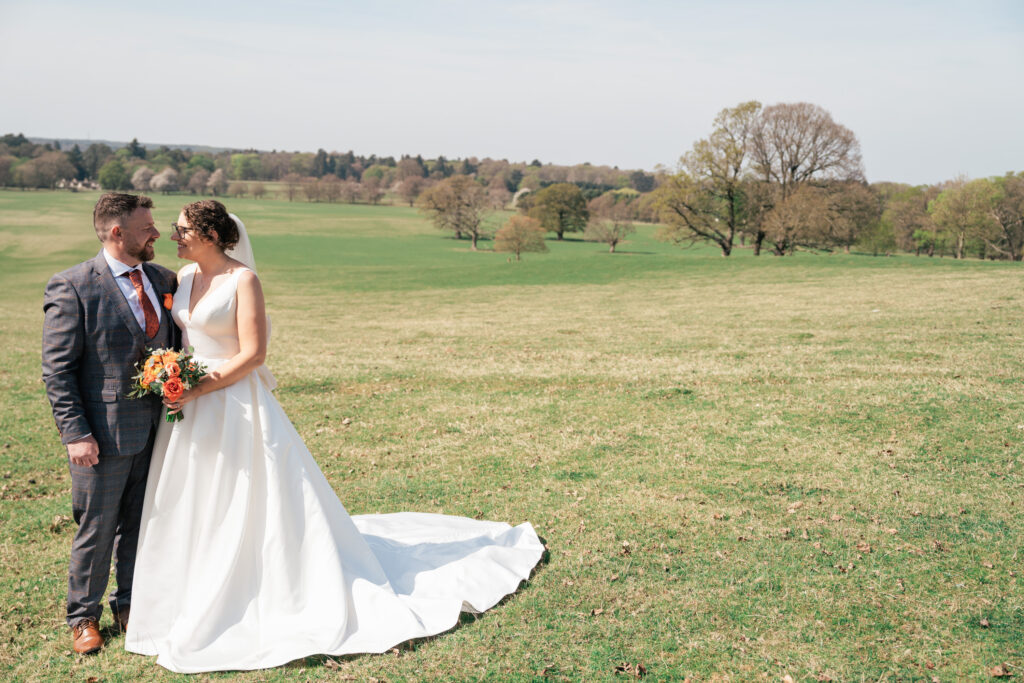 A bride in a white dress and a groom in a checked suit stand together on a grassy field, holding a bouquet and gazing at each other, with trees and open sky in the background, perfectly captured by a Surrey wedding photographer.