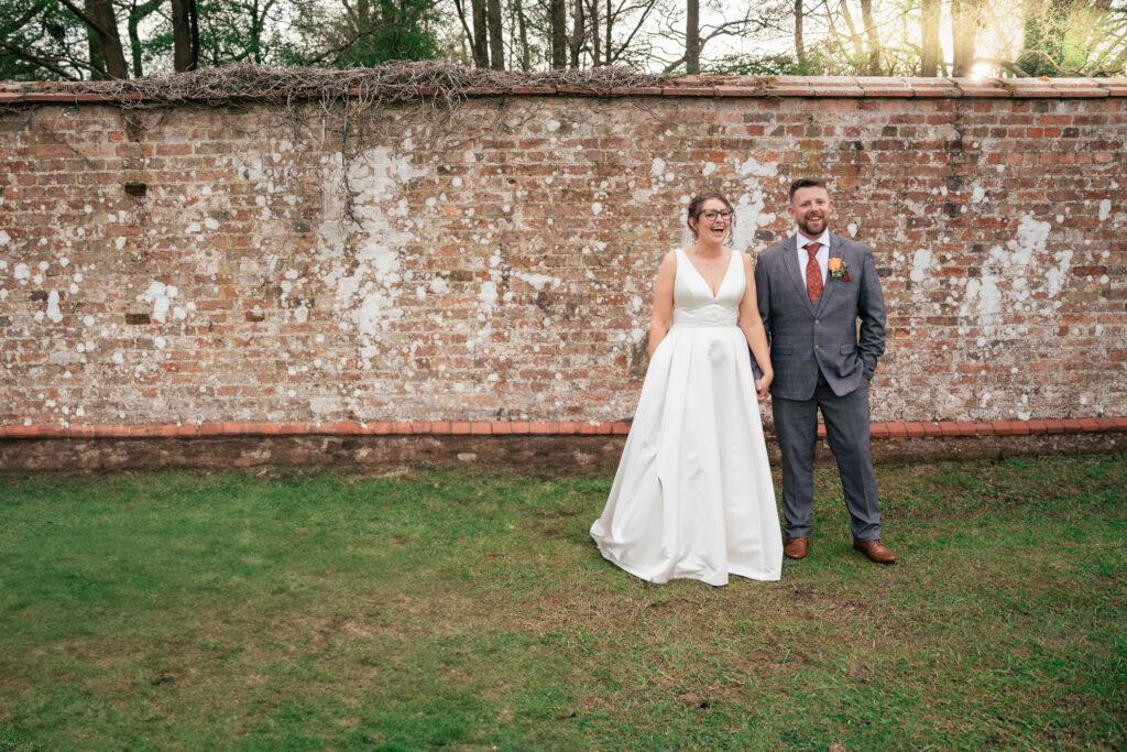 A bride in a white gown and a groom in a gray suit stand smiling and holding hands on a grassy lawn, captured beautifully by a Surrey wedding photographer in front of a weathered brick wall outdoors.