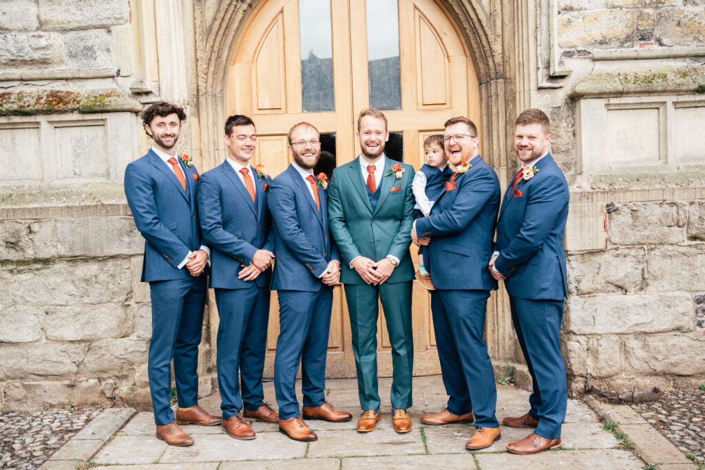 Six men in suits, one holding a small child, stand smiling in front of a large wooden door set in a stone building, likely at a wedding or formal event. Everyone appears happy and well-dressed.