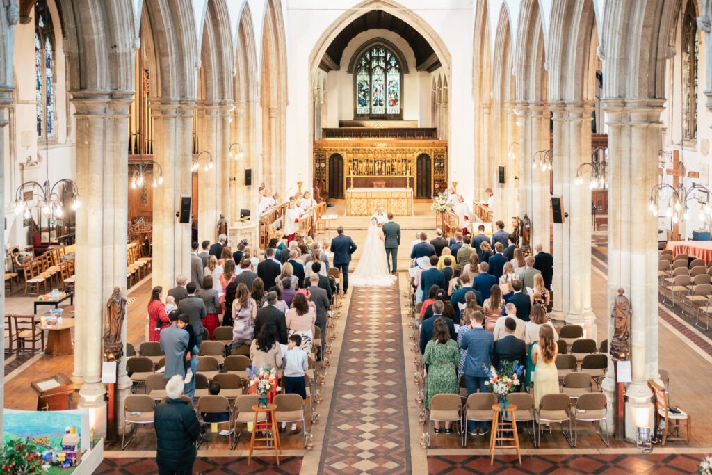 A bride and groom stand at the altar inside a large, bright church filled with seated guests, tall columns, and stained glass windows. The wedding ceremony is in progress, viewed from the back of the aisle.