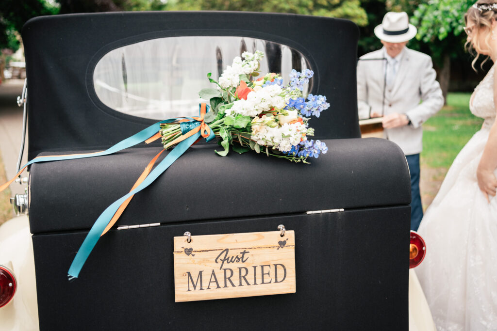 A bouquet of flowers and a Just Married sign decorate the back of a vintage car; a person in a light suit and hat stands nearby, while a woman in a wedding dress is partly visible on the right.