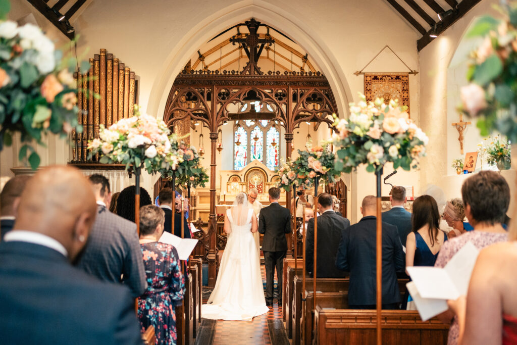 A bride and groom stand at the altar in a church during their wedding ceremony, surrounded by guests seated in pews. Captured by a Surrey wedding photographer, the church is decorated with flowers and boasts a stained-glass window above the altar.