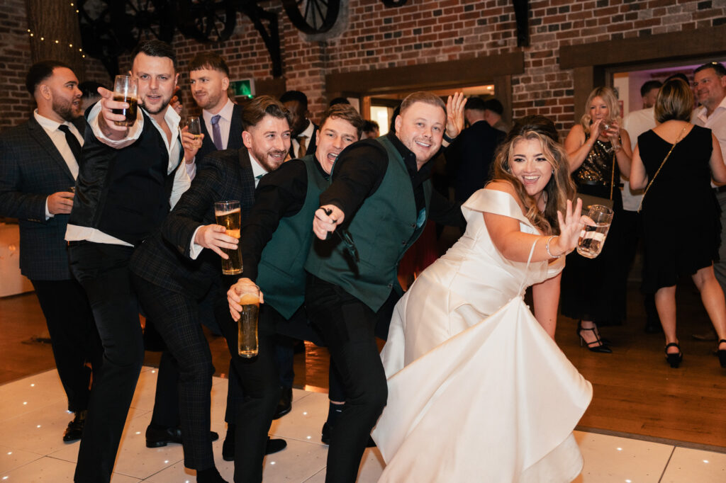 A group of people, including a bride in a white dress, pose playfully on a dance floor at a wedding reception, smiling and holding drinks, as captured by a Surrey wedding photographer with other guests and brick walls in the background.