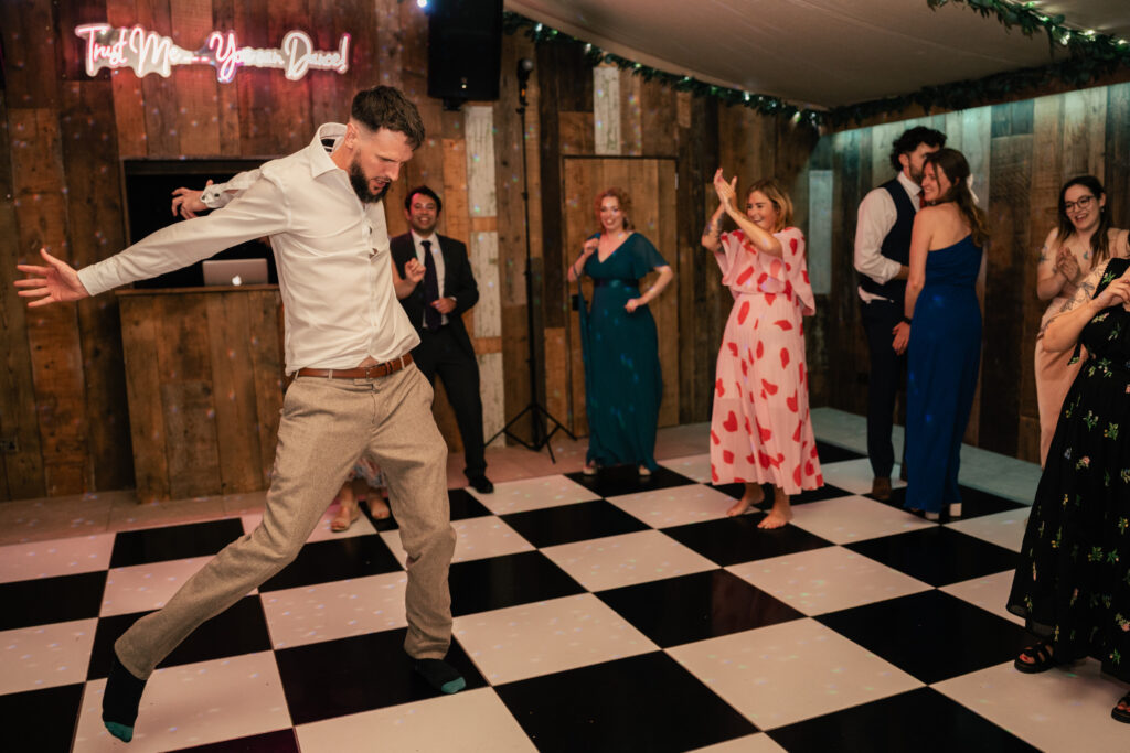 A man joyfully dances on a black-and-white checkered floor at a party, while other people in formal attire watch, smile, and join in on a warmly lit dance floor with rustic wooden walls.