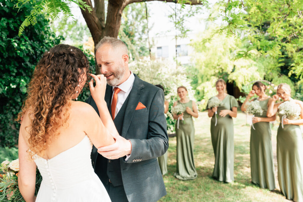 A bride in a white dress wipes tears from her eyes while holding hands with a man in a suit. Four bridesmaids in green dresses stand behind them outside, smiling and holding bouquets.