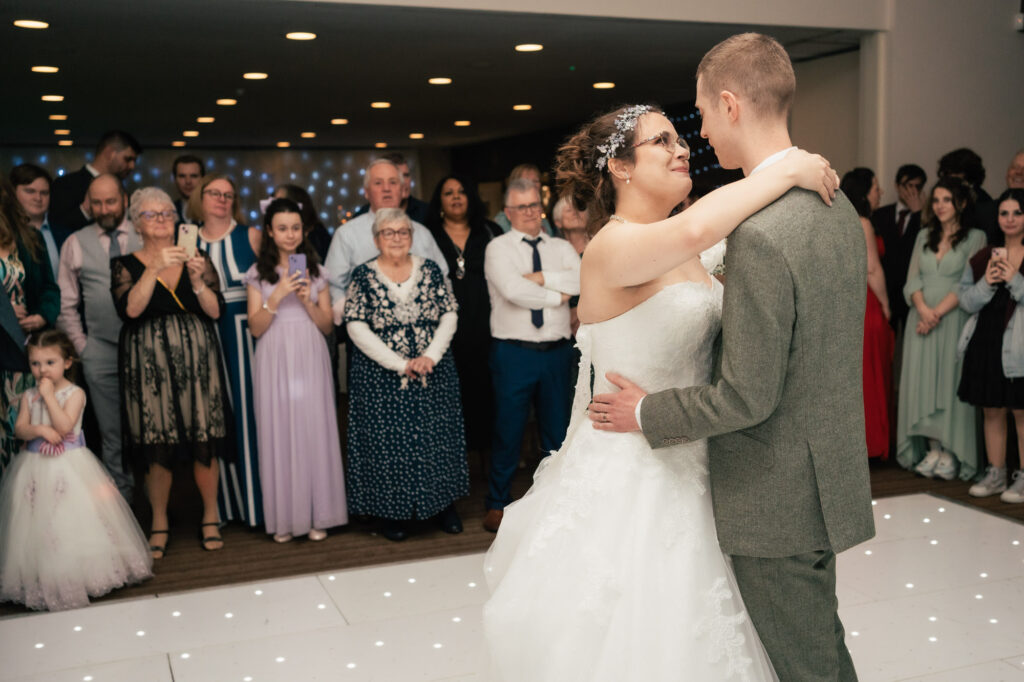 A bride and groom share their first dance at a wedding reception, surrounded by smiling guests. Captured by a Surrey wedding photographer, the couple shines on the lit white dance floor as friends and family watch and take photos.