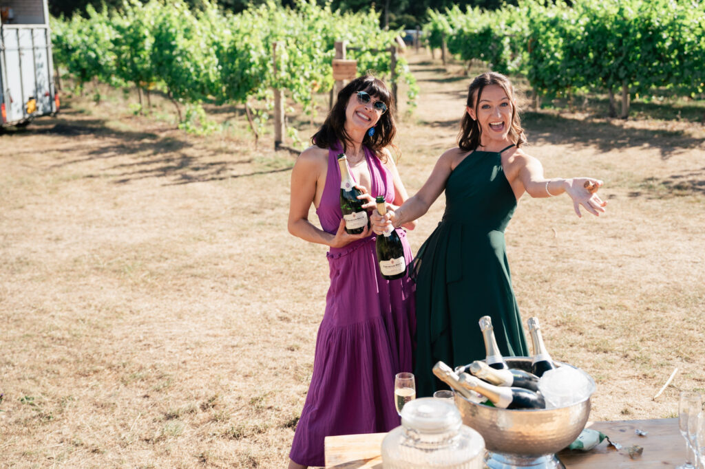 Two women in dresses, one purple and one green, smile and pose holding wine bottles outdoors in a sunny vineyard next to a table with an ice bucket and drinks. Grapevines are visible in the background.