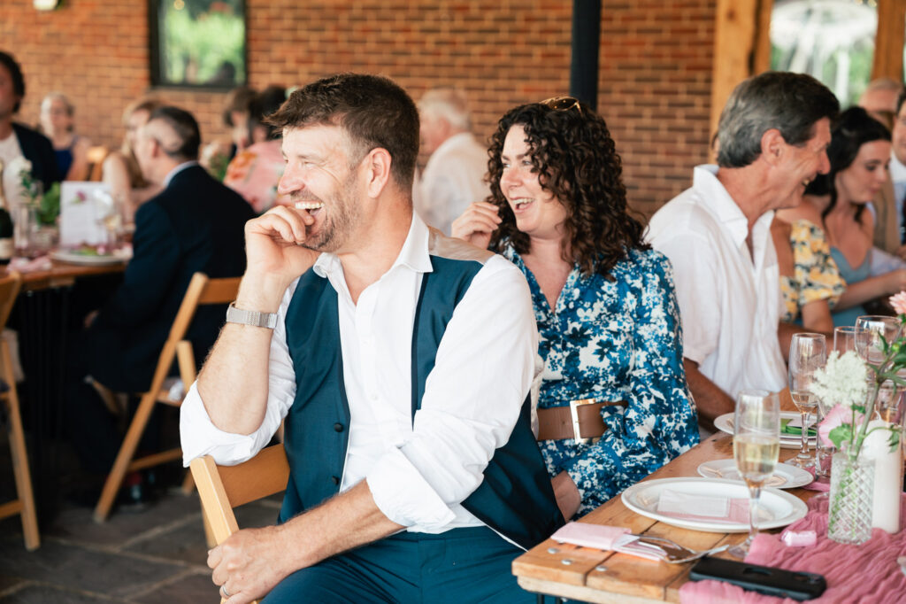 A man in a white shirt and vest laughs while sitting at a table with other guests at an indoor event. People around him are smiling, and the atmosphere appears joyful and relaxed.