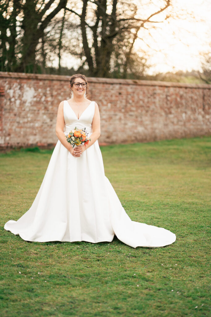 A bride in a white sleeveless wedding dress stands on grass, holding a bouquet of flowers. Captured by a Surrey wedding photographer, she poses before a brick wall and trees with bare branches in the gentle, late daylight.