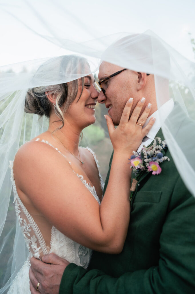 A bride and groom stand close under a sheer veil, smiling lovingly at each other. The bride gently holds the groom’s face, and both look joyful and intimate on their wedding day. The groom wears glasses and a green suit.
