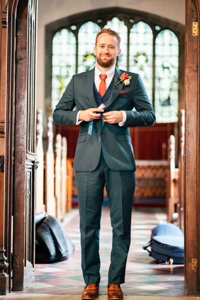 A man in a teal suit and brown shoes stands in a church doorway, buttoning his jacket and smiling. He wears a red tie and boutonnière, with light streaming through stained glass windows behind him.